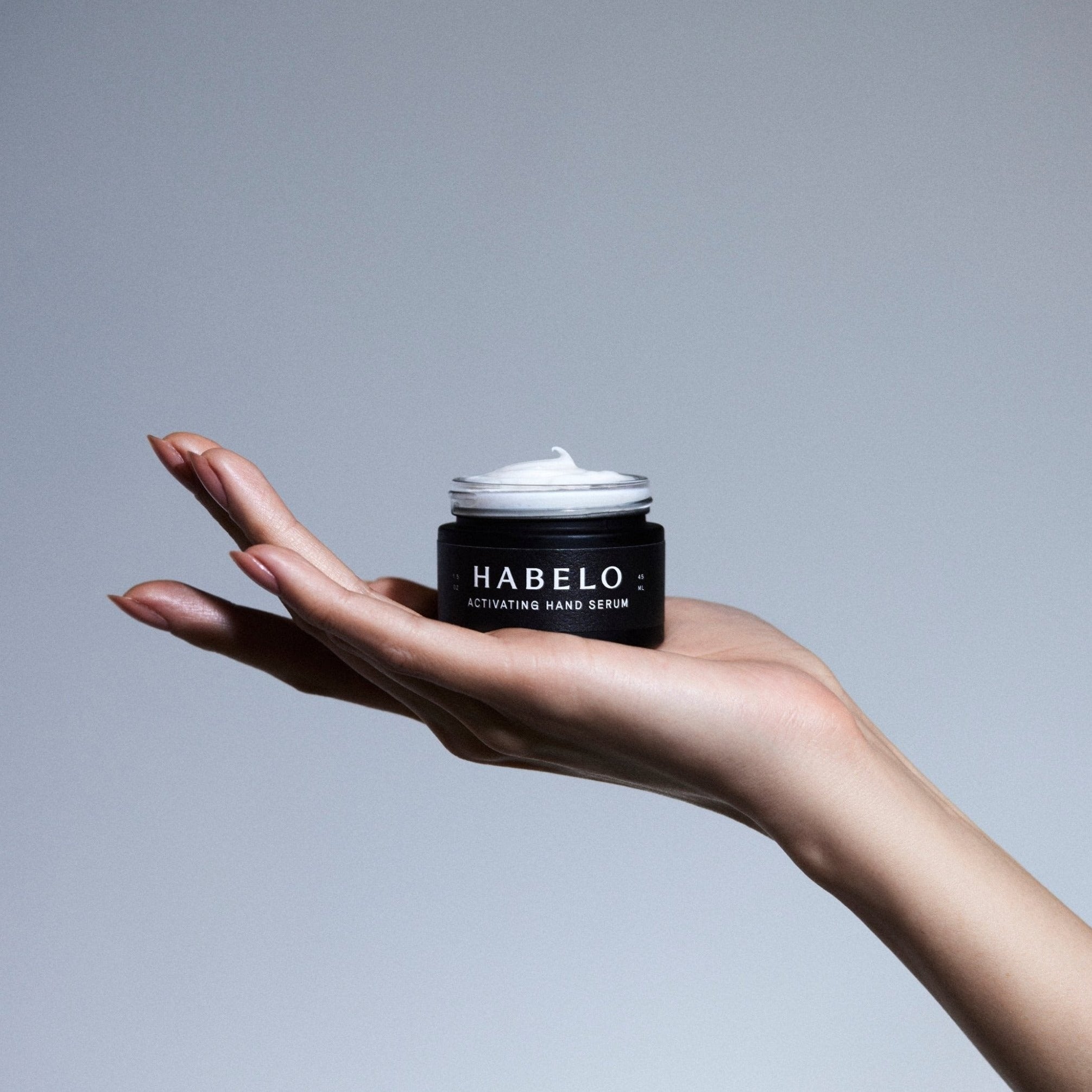 A hand holding an open jar of Habelo Activating Hand Serum